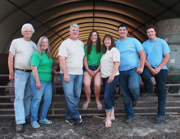 Family of farmers posing together inside a barn, showcasing their commitment to agriculture and livestock care. They wear matching colored shirts, representing teamwork and family values. The background features a rustic barn environment, emphasizing a connection to farming life.